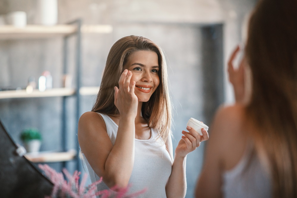 Pandemic self care girl applying moisturizing cream on her face near mirror indoors